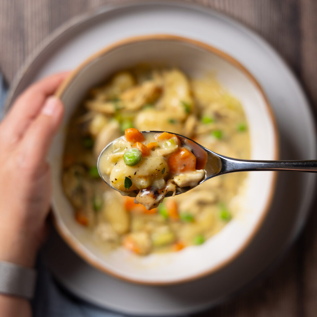 A bowl sits on a plate, inside the bowl is a dish called Chicken Pot Pie Gnocchi; carrots, celery and mushrooms in a creamy sauce with gnocchi. A spoon holds some of the food. A napkin is nearby.