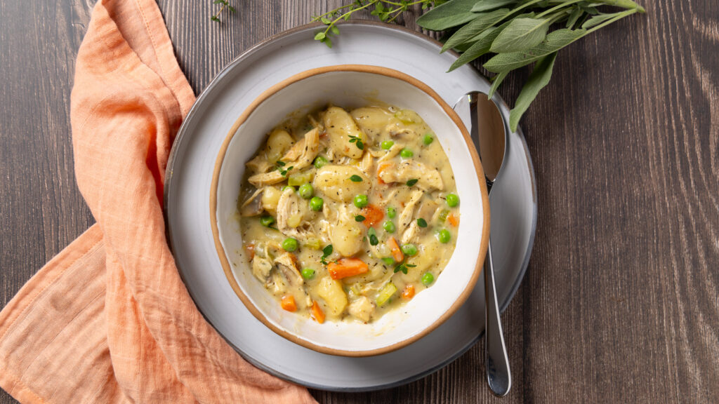 A bowl sits on a plate, inside the bowl is a dish called Chicken Pot Pie Gnocchi; carrots, celery and mushrooms in a creamy sauce with gnocchi. Some herbs are near the plate. A napkin is nearby.