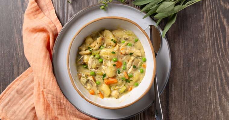 A bowl sits on a plate, inside the bowl is a dish called Chicken Pot Pie Gnocchi; carrots, celery and mushrooms in a creamy sauce with gnocchi. Some herbs are near the plate. A napkin is nearby.