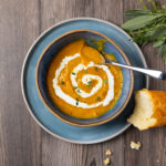 Butternut squash soup with a swirl of yogurt in a bowl. Fresh herbs and a piece of crusty bread are next to the bowl.