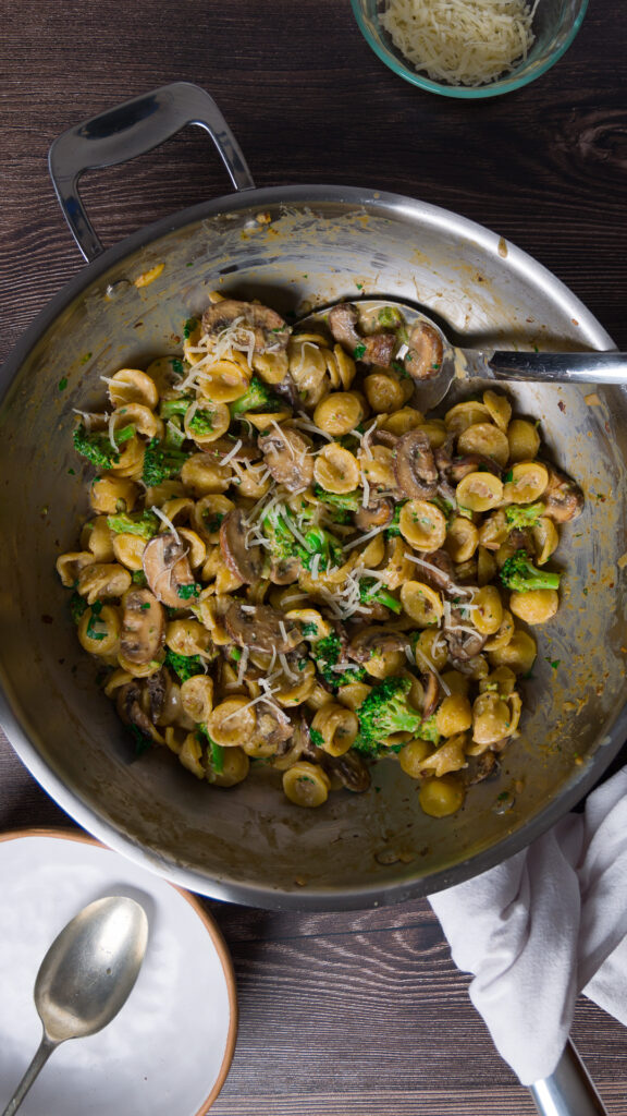 A large skillet holds a pasta dish with mushrooms, broccoli, and a creamy sauce.