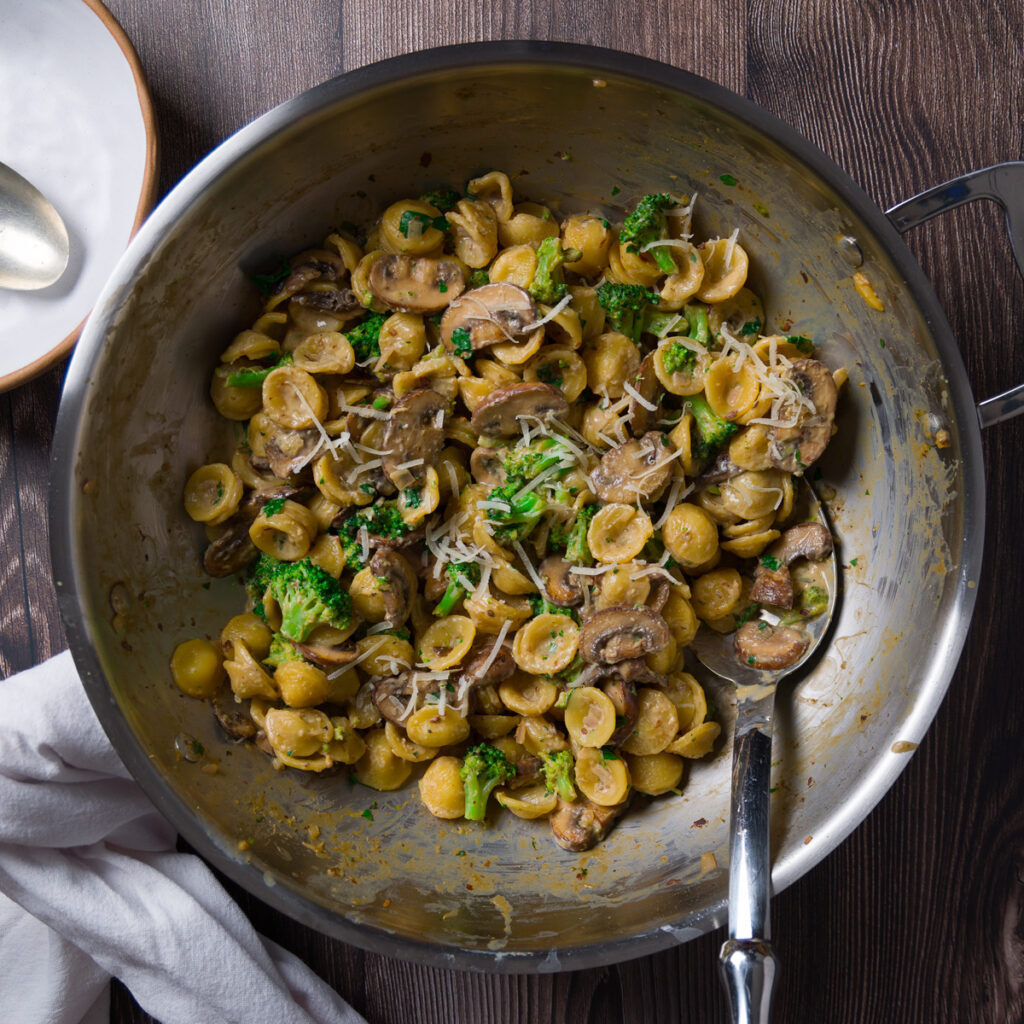 A large skillet holds a pasta dish with mushrooms, broccoli, and a creamy sauce.