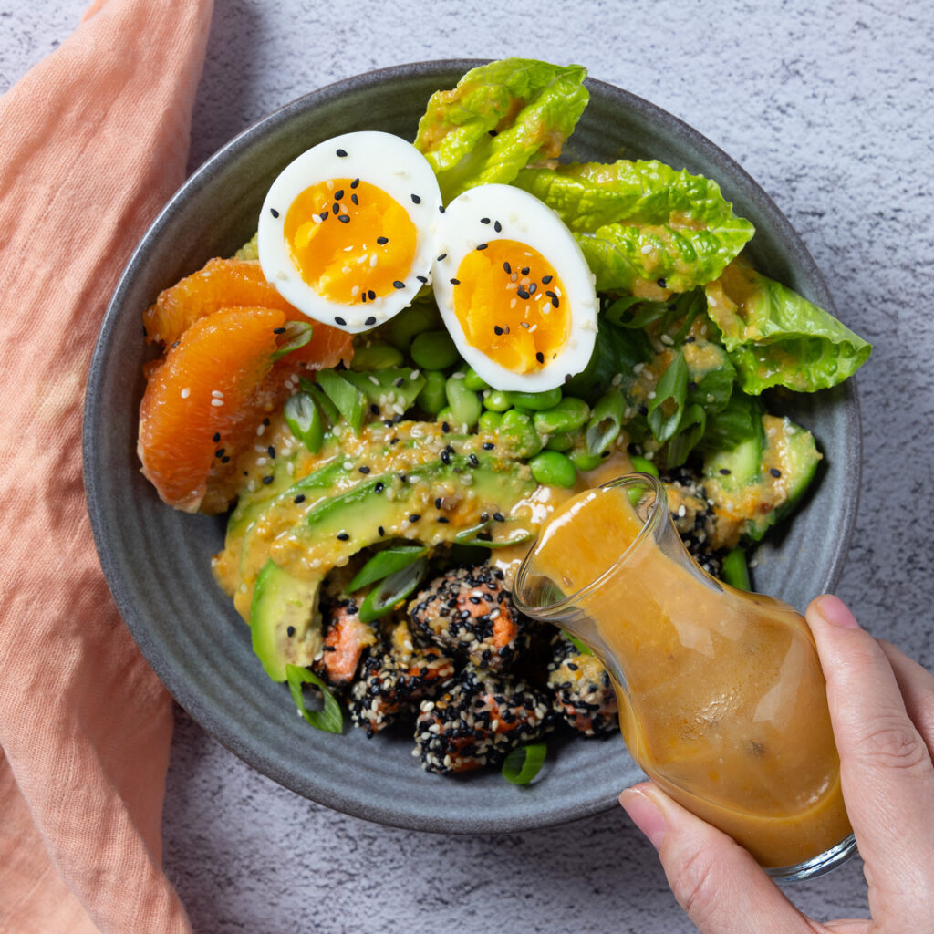 A bowl containing sesame crusted salmon chunks, orange segments, avocado slides, lettuce leaves, edamame beans. A carafe of dressing. Chopsticks rest on the bowl and a pink napkin is next to the bowl.