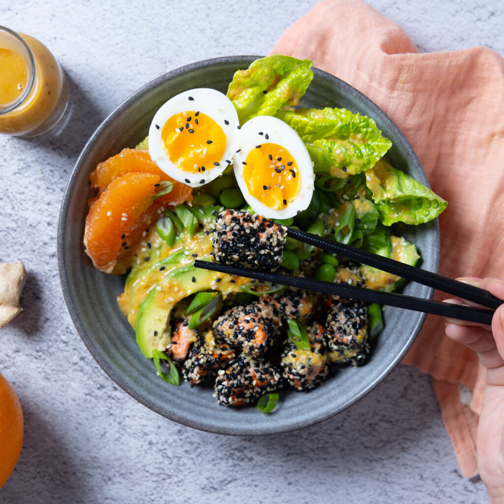 A bowl containing sesame crusted salmon chunks, orange segments, avocado slides, lettuce leaves, edamame beans. A carafe of dressing. Chopsticks rest on the bowl and a pink napkin is next to the bowl.