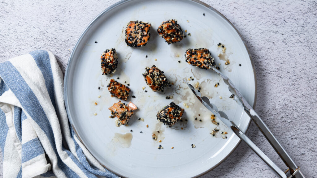 A plate contains freshly seared sesame seed-crusted salmon chunks. Tongs rest on the plate.