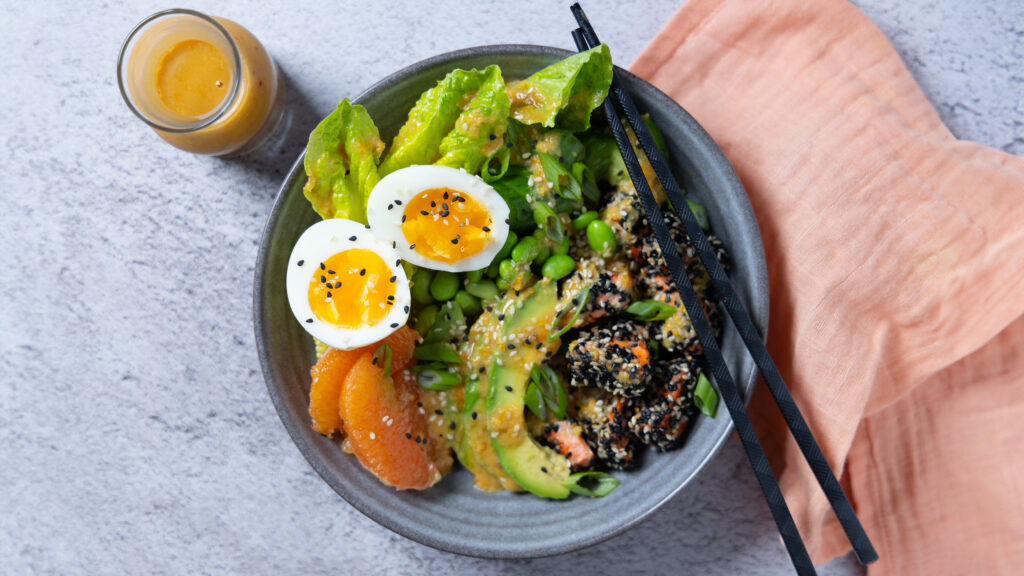 A bowl containing sesame crusted salmon chunks, orange segments, avocado slides, lettuce leaves, edamame beans. A carafe of dressing. Chopsticks rest on the bowl and a pink napkin is next to the bowl.