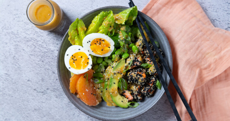 A bowl containing sesame crusted salmon chunks, orange segments, avocado slides, lettuce leaves, edamame beans. A carafe of dressing. Chopsticks rest on the bowl and a pink napkin is next to the bowl.