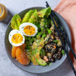 A bowl containing sesame crusted salmon chunks, orange segments, avocado slides, lettuce leaves, edamame beans. A carafe of dressing. Chopsticks rest on the bowl and a pink napkin is next to the bowl.