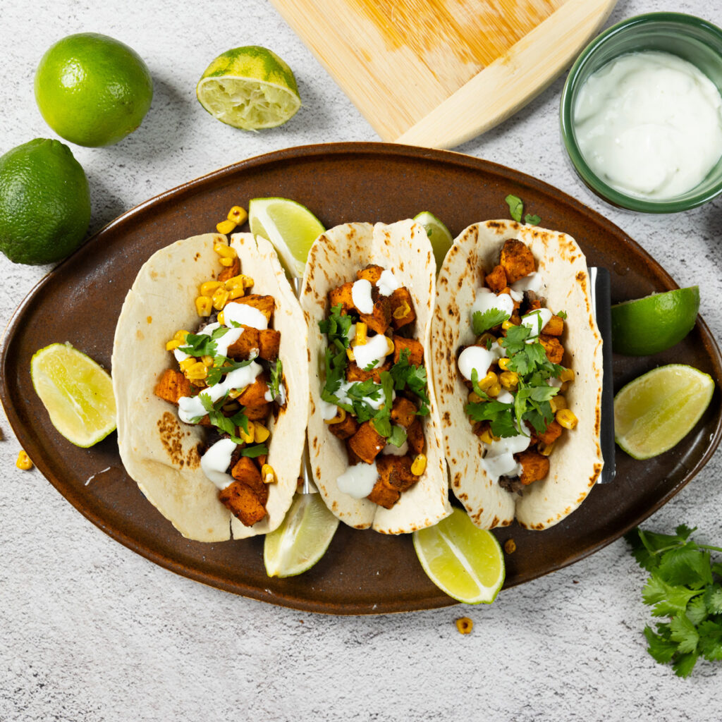 A trio of sweet potato tacos, with refried black beans, charred corn, soft wheat tortilla shells, lime crema, chopped cilantro, surrounded by limes.