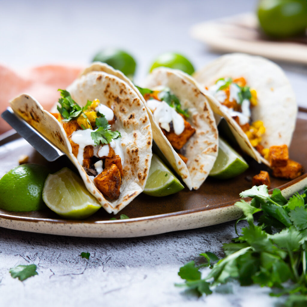 A trio of sweet potato tacos, with refried black beans, charred corn, soft wheat tortilla shells, lime crema, chopped cilantro.