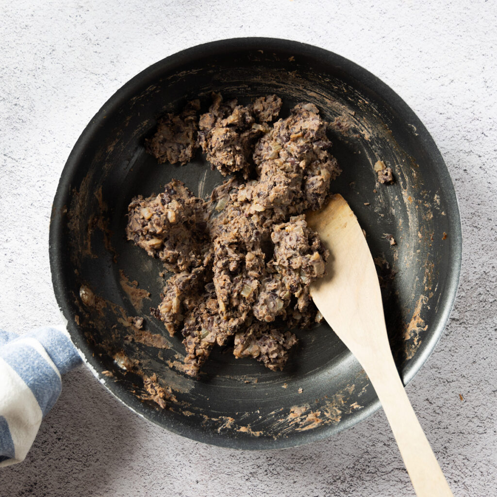 A skillet with refried black beans. A wooden spatula is in the pan.