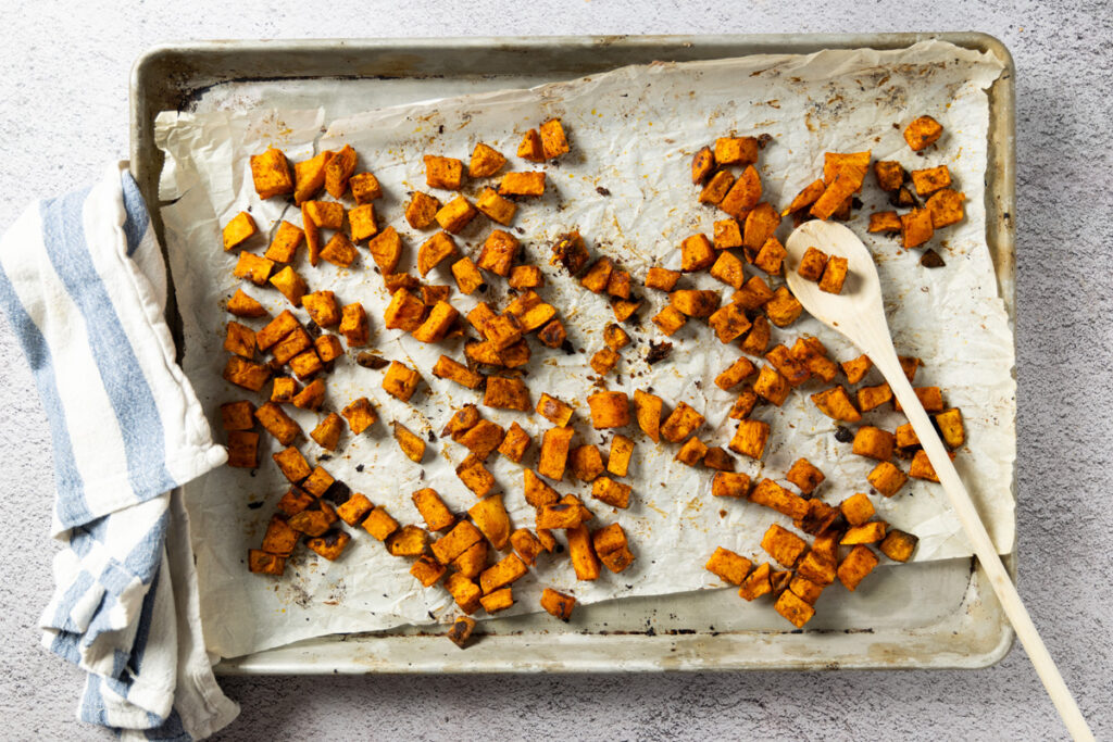 A sheet pan, lined with parchment paper, holds cubed, roasted sweet potato.