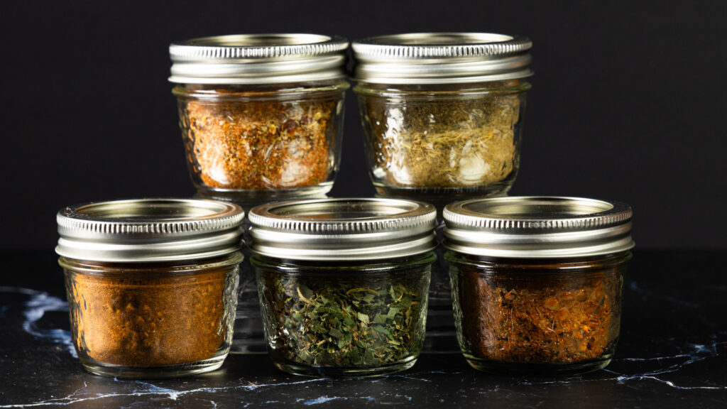 Five jars of homemade spices, containing taco seasoning, cajun seasoning, italian seasoning, poultry seasoning, and pumpkin spice.