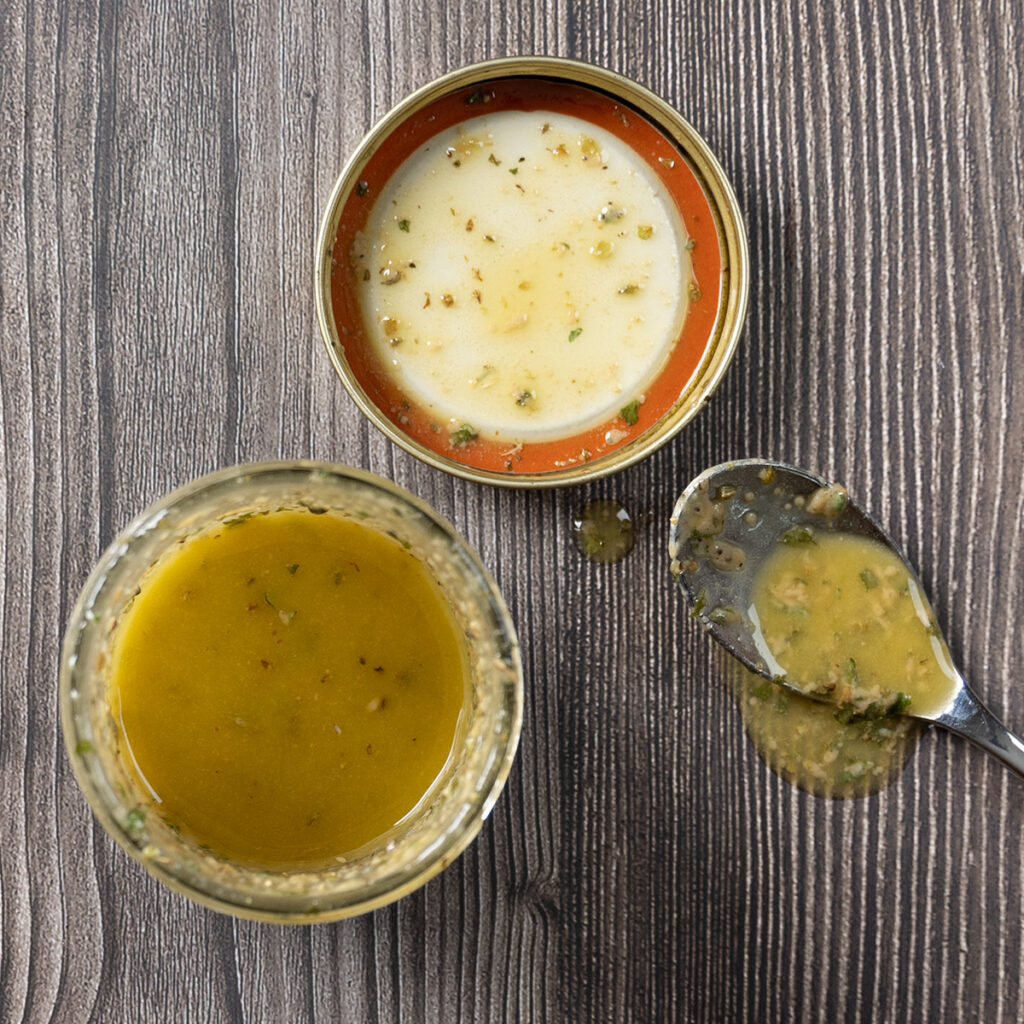 A jar containing homemade Greek dressing, the lid and a spoon sit nearby.