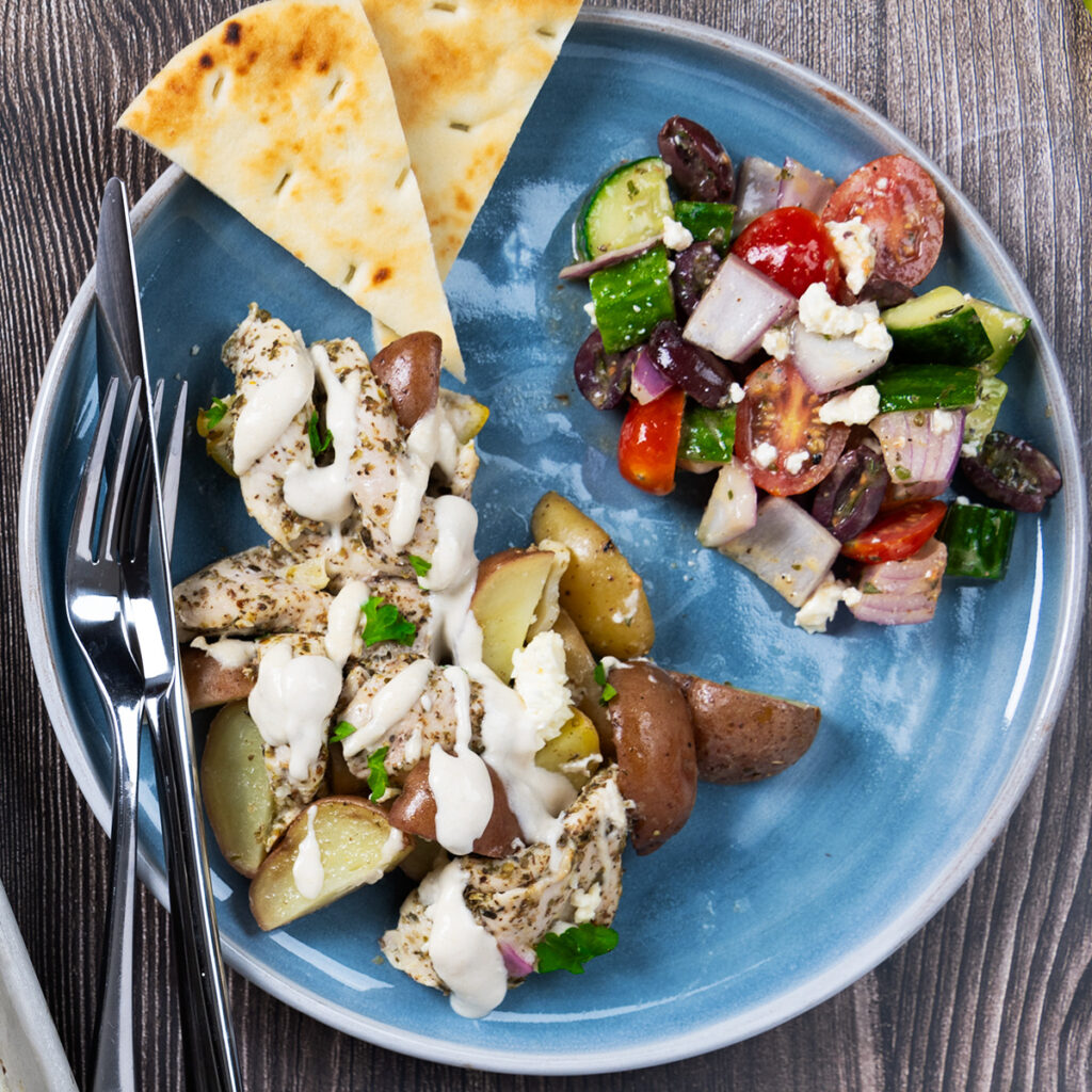 A plate with chicken and potatoes, topped with a tahini sauce, next to a greek salad and pita bread.