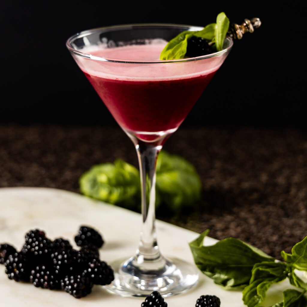 A cocktail sour, containing blackberries, basil, and gin is in a martini glass which is on a marble tray. The glass is surrounded by fresh blackberries and basil leaves.