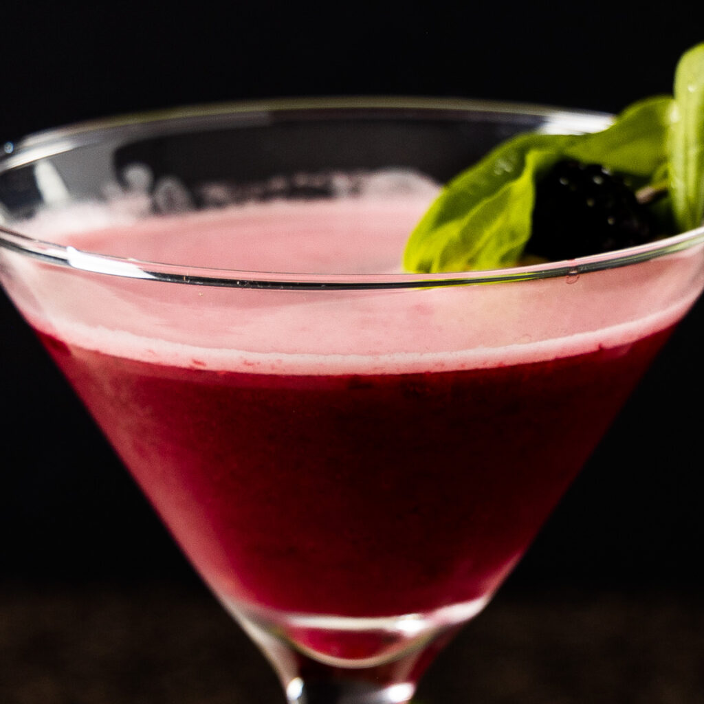 A cocktail sour, containing blackberries, basil, and gin is in a martini glass which is on a marble tray. The glass is surrounded by fresh blackberries and basil leaves.