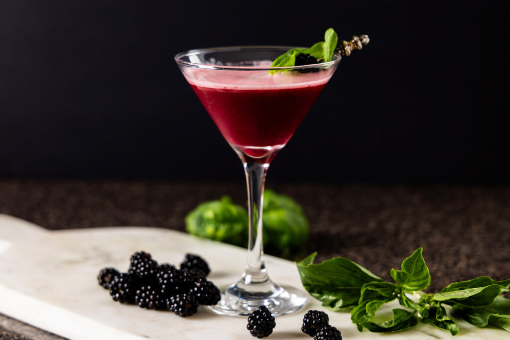 A cocktail sour, containing blackberries, basil, and gin is in a martini glass which is on a marble tray. The glass is surrounded by fresh blackberries and basil leaves.