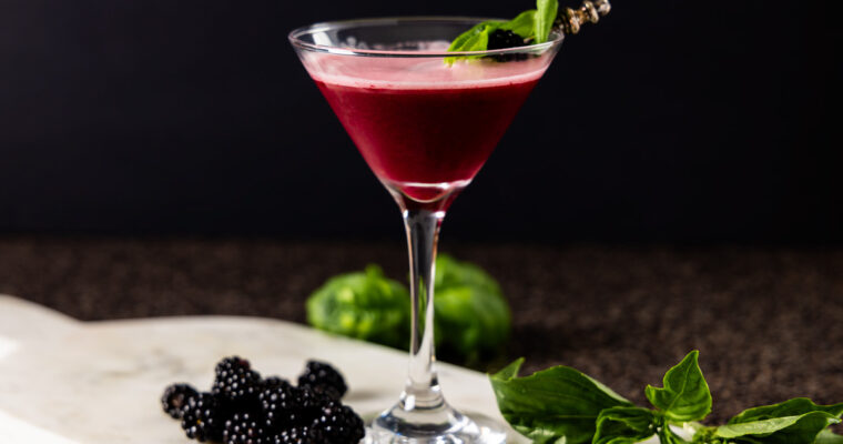 A cocktail sour, containing blackberries, basil, and gin is in a martini glass which is on a marble tray. The glass is surrounded by fresh blackberries and basil leaves.