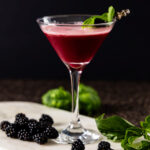 A cocktail sour, containing blackberries, basil, and gin is in a martini glass which is on a marble tray. The glass is surrounded by fresh blackberries and basil leaves.