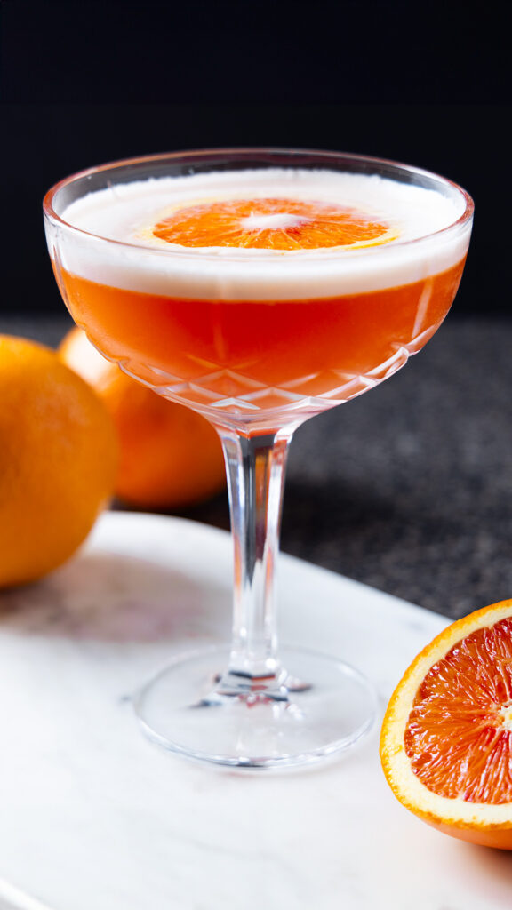 A negroni sour in a cocktail glass, with various oranges around.
