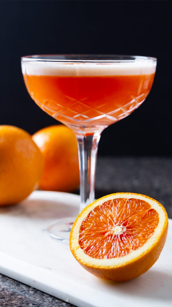 A negroni sour in a cocktail glass, with various oranges around.
