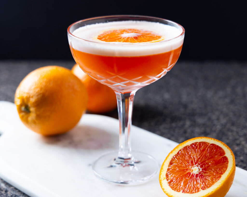 A negroni sour in a cocktail glass, with various oranges around.
