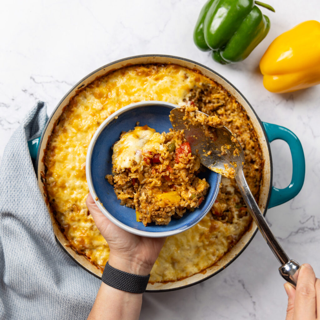 Photo of a skillet, filled with Stuffed Pepper Casserole. A large metal spoon is scooping out a serving of the dish into a bowl; a green and yellow bell pepper sit nearby. A towel is wrapped around the handle of the skillet.