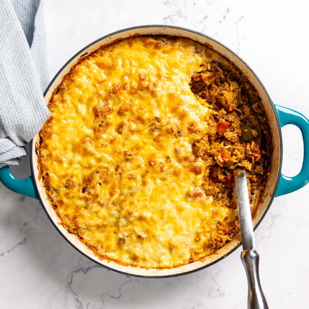 Photo of a skillet, filled with Stuffed Pepper Casserole. A large metal spoon is scooping out a serving of the dish; a green and yellow bell pepper sit nearby. A towel is wrapped around the handle of the skillet.