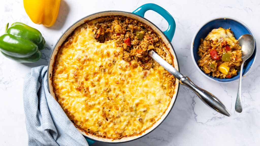 Photo of a skillet, filled with Stuffed Pepper Casserole. A large metal spoon sits in the skillet with the casserole; a green and yellow bell pepper sit nearby and a bowl with a serving of the rice dish sits next to the skillet. A towel is wrapped around the handle of the skillet.