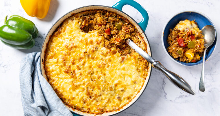 Photo of a skillet, filled with Stuffed Pepper Casserole. A large metal spoon sits in the skillet with the casserole; a green and yellow bell pepper sit nearby and a bowl with a serving of the rice dish sits next to the skillet. A towel is wrapped around the handle of the skillet.