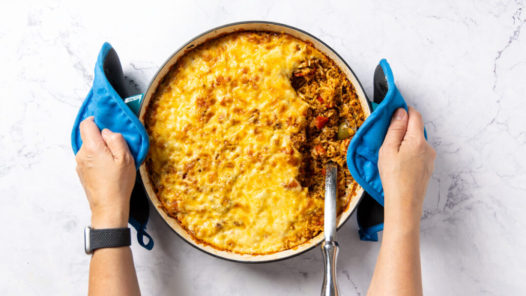 Photo of a skillet, filled with Stuffed Pepper Casserole. A large metal spoon is in the casserole; a green and yellow bell pepper sit nearby. A towel is wrapped around the handle of the skillet. Hands with oven mitts are holding the handles of the skillet.