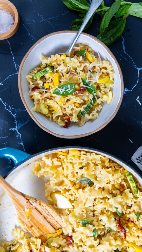 summer squash pasta in a large skillet, with basil, lemon, red peppers, with a bowl of pasta with a spoon nearby.