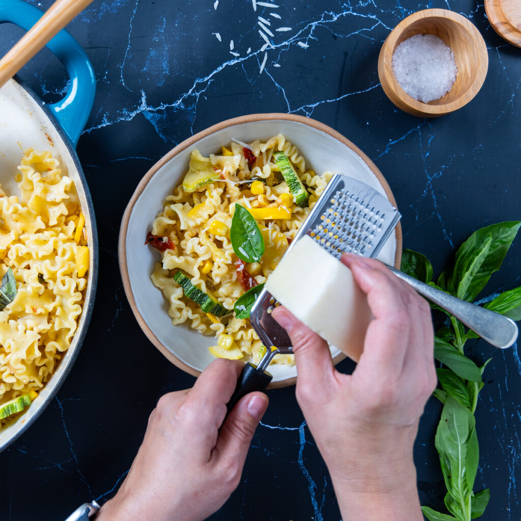 Hands are grating cheese over top a summer squash pasta in a large skillet, with basil, lemon, red peppers.