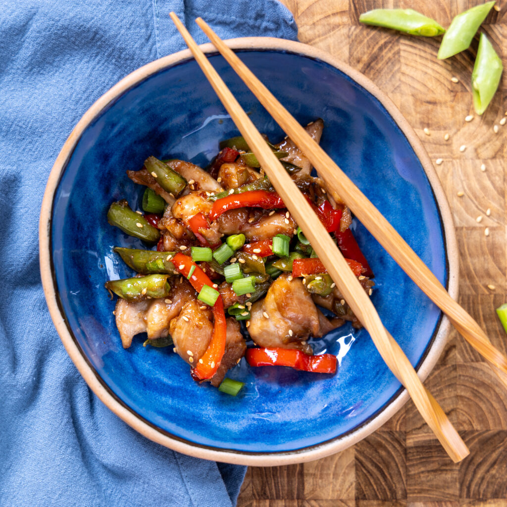 A blue bowl contains chili garlic chicken. Chopsticks rest on one edge of the bowl. A blue napkin site under the bowl. Chopped pea pods are next to the bowl.