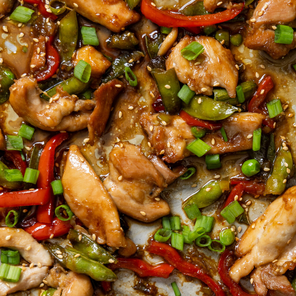 A close up photo of food, with chicken pieces, sugar snap peas, red bell peppers, green onions, sesame seeds, in a chili garlic sauce.