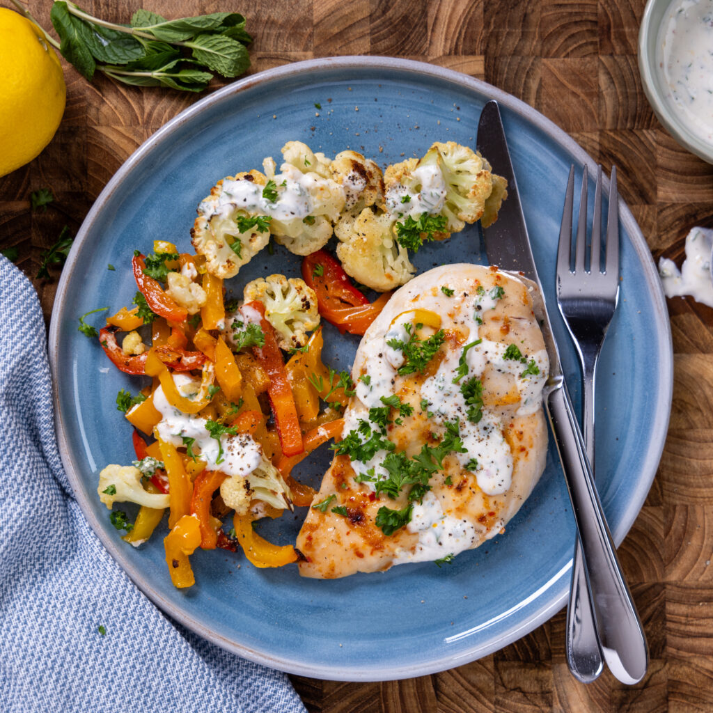 A blue plate has harissa-spiced chicken breast, roasted vegetables, drizzled with a herbed yogurt sauce.