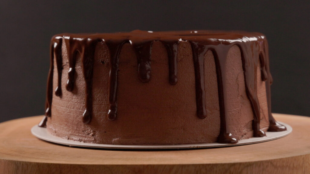 A chocolate layer cake with ganache drip sits on a wooden tray.