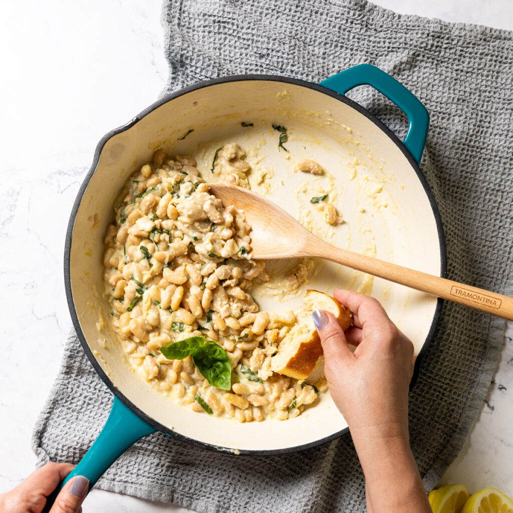 creamy lemon parmesan beans in a skillet, a hand is using a chunk of crusty bread to mop up every delicious drop of the amazing sauce
