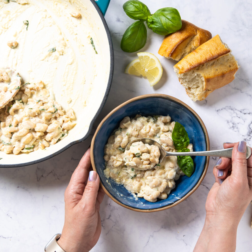 Creamy lemon parmesan beans in a bowl; a hand holding a spoon is taking a scoop of beans from the bowl. A skillet used to cook beans is next to the bowl.