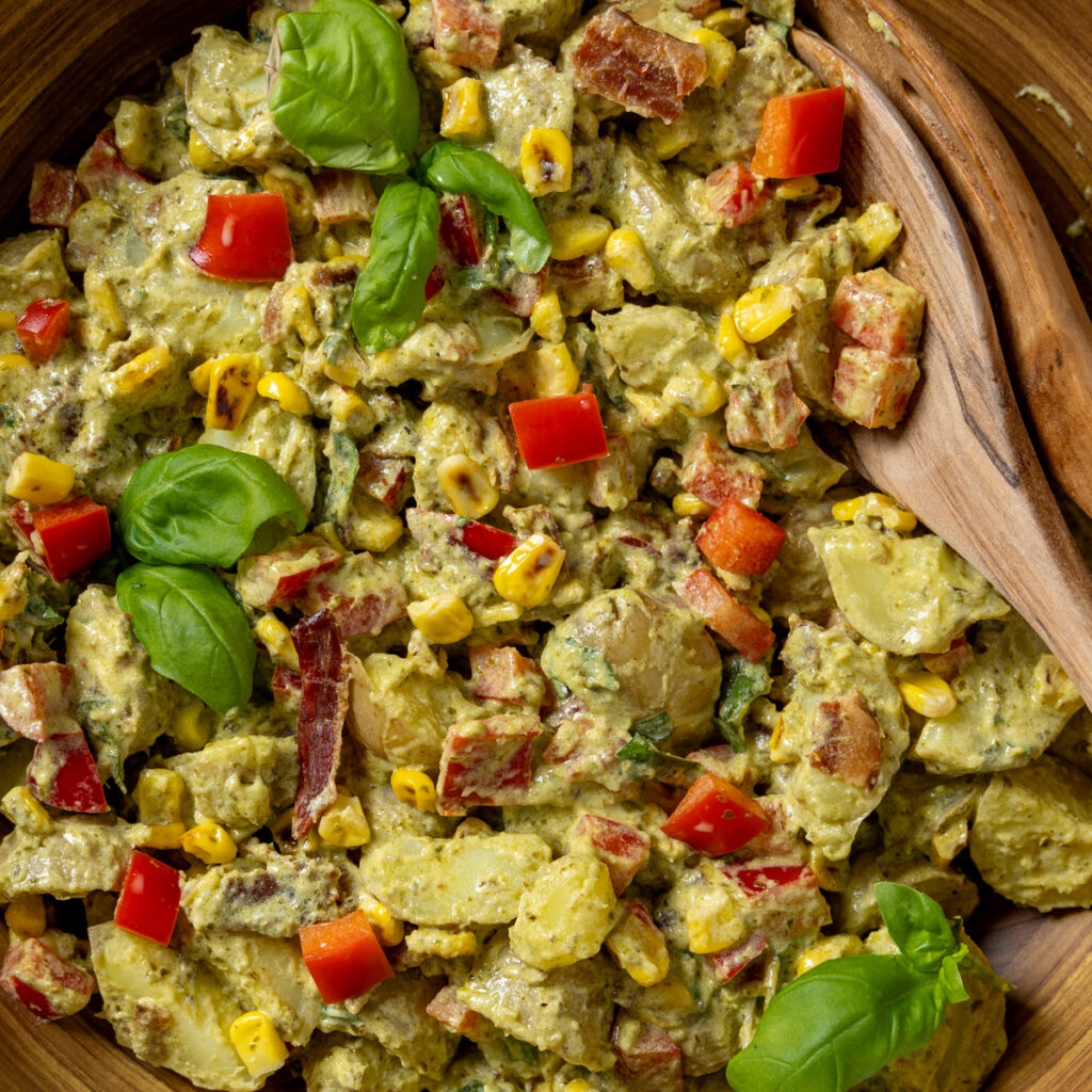 Creamy pesto potato salad with corn, bacon, and basil leaves in a bowl with serving spoons. The bowl sits on a red and white tea towel.