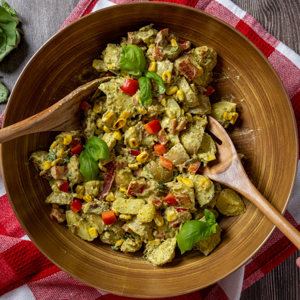 Creamy pesto potato salad with corn, bacon, and basil leaves in a bowl with serving spoons. The bowl sits on a red and white tea towel.