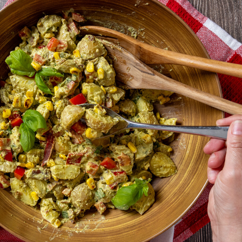 Creamy pesto potato salad with corn, bacon, and basil leaves in a bowl with serving spoons. The bowl sits on a red and white tea towel.