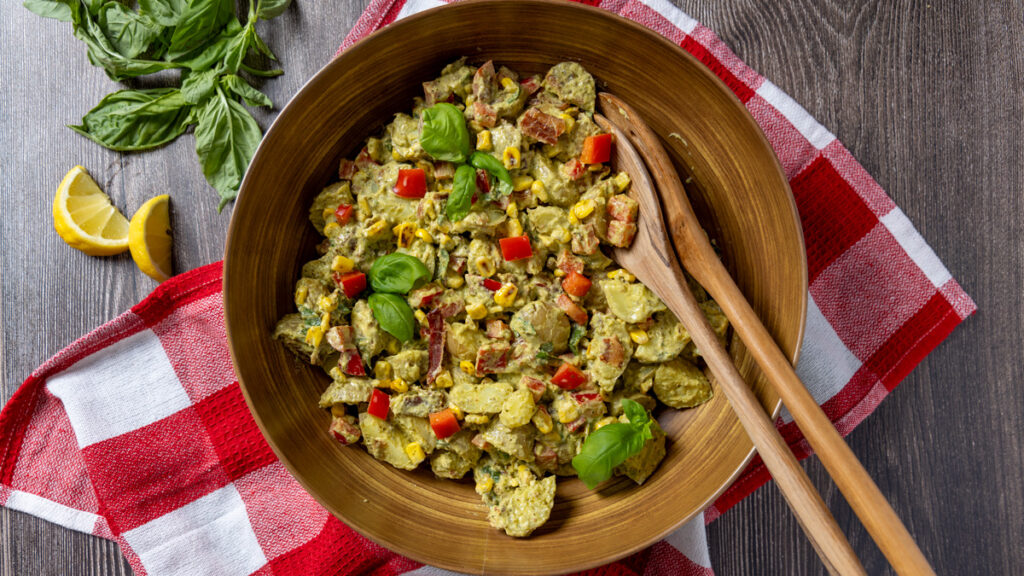 Creamy pesto potato salad with corn, bacon, and basil leaves in a bowl with serving spoons. The bowl sits on a red and white tea towel.