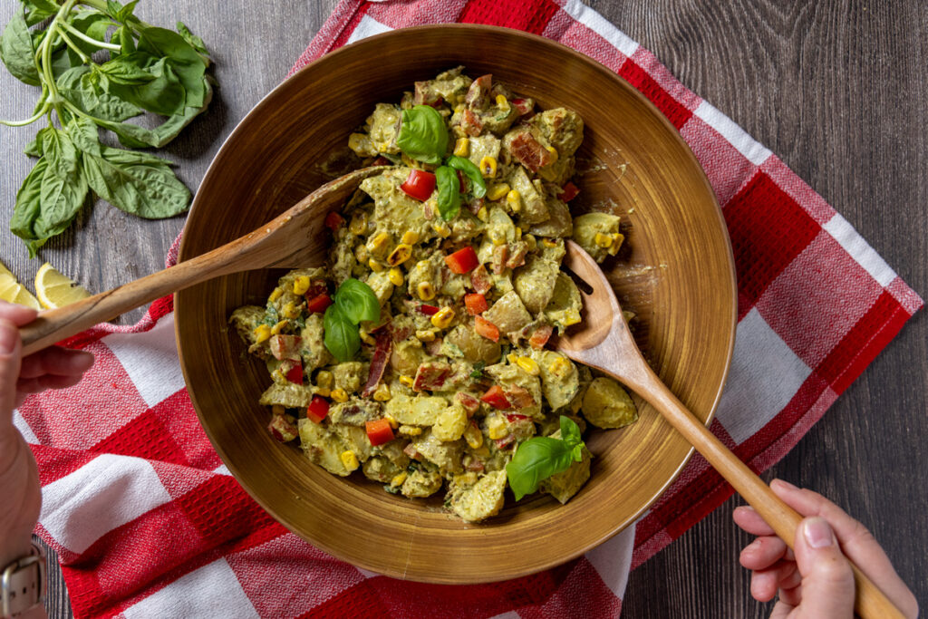 Creamy pesto potato salad with corn, bacon, and basil leaves in a bowl with serving spoons. The bowl sits on a red and white tea towel.