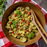 Creamy pesto potato salad with corn, bacon, and basil leaves in a bowl with serving spoons. The bowl sits on a red and white tea towel.